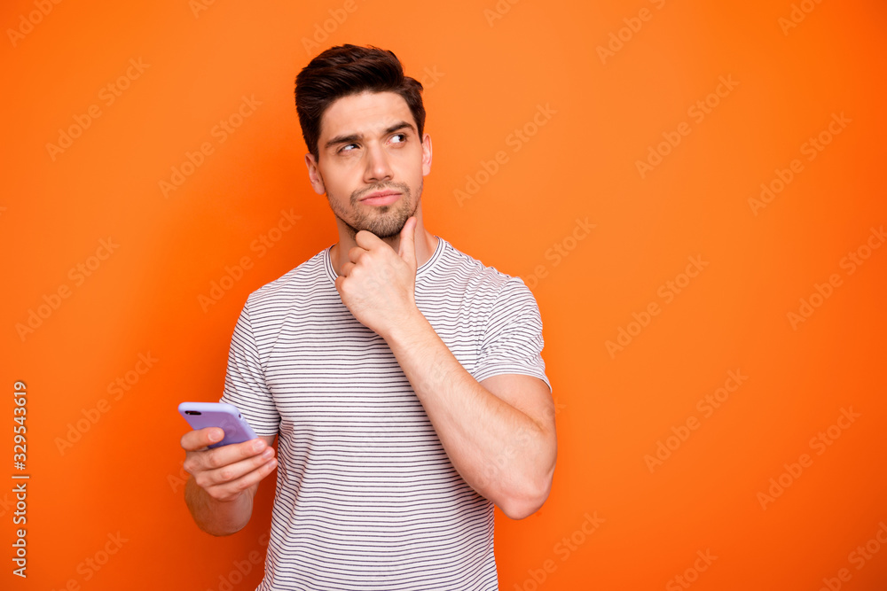 Photo of minded serious millennial guy hold telephone deep think new instagram blog post text arm on chin look side wear striped t-shirt isolated bright orange color background - obrazy, fototapety, plakaty 