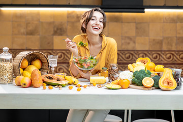 Wall Mural - Portrait of a young cheerful woman eating salad at the table full of healthy raw vegetables and fruits on the kitchen at home. Concept of vegetarianism, healthy eating and wellness