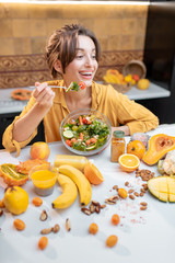 Wall Mural - Portrait of a young cheerful woman eating salad at the table full of healthy raw vegetables and fruits on the kitchen at home. Concept of vegetarianism, healthy eating and wellness