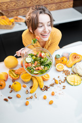 Wall Mural - Portrait of a young cheerful woman eating salad at the table full of healthy raw vegetables and fruits on the kitchen at home. Concept of vegetarianism, healthy eating and wellness