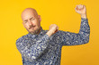 © Augustino - Fabulous at any age. Portrait of 40-year-old dancing man feeling happy over light yellow background in blue shirt. Funny face. Hipster style. Bald shaved head. Studio shot