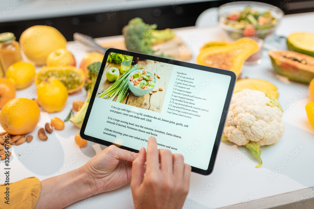 Woman looking on the digital recipe, using touchscreen tablet while cooking healthy meal on the kitchen at home, close-up view on the screen - obrazy, fototapety, plakaty 
