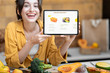 © rh2010 - Woman holding digital tablet with launched online shopping market while standing on the kitchen with lots of fresh food on the table
