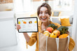 © rh2010 - Happy woman holding a digital tablet with launched online store while standing with shopping bag full of fresh products at home. Concept of buying online using mobile devices