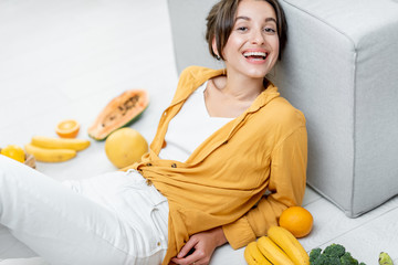 Wall Mural - Portrait of a young and cheerful woman sitting with lots of fresh fruits and vegetables on the floor at home. Photo carried in yellow color. Concept of wellbeing, healthy food and homeliness