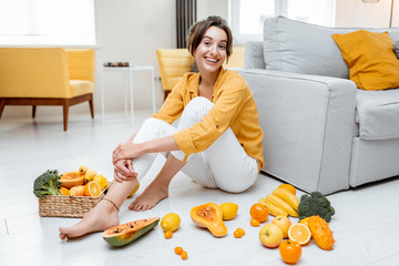 Wall Mural - Portrait of a young and cheerful woman sitting with lots of fresh fruits and vegetables on the floor at home. Photo carried in yellow color. Concept of wellbeing, healthy food and homeliness