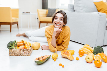 Wall Mural - Portrait of a young and cheerful woman lying with lots of fresh fruits and vegetables on the floor at home. Photo carried in yellow color. Concept of wellbeing, healthy food and homeliness