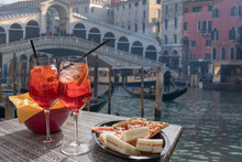 Cocktail, Aperitif For Two With The View Of Venice In The Background. Two Glasses Of Spritz With Lemon And Straws And A Plate Of Snacks