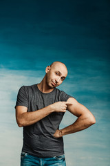 Young bald man in a gray T-shirt and jeans boasts of his biceps on a blue background.