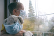 Leinwandbild Motiv Sad illness child on home quarantine. Boy and his teddy bear both in protective medical masks sits on windowsill and looks out window. Virus protection, coronavirus pandemic, prevention epidemic.