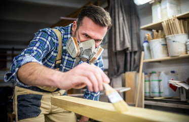 Carpenter with mask applies paint using paintbrush in carpentry workshop