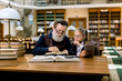 © sofiko14 - Senior man teacher professor teaching little girl, showing her a book, while sitting in the library with vintage bookcases background. Girl holds tablet and looking at the book