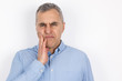 © studioprodakshn - adult handsome man with grey hair wearing blue shirt touching cheek with hand suffering from sudden toothache standing on isolated white