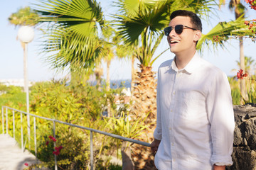 A man in a white shirt and dark glasses on a background of palm trees has a warm day. The guy is waiting. A man in the tropics enjoys life, vacation alone. The man is resting alone.