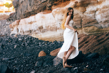 Wall Mural - Gorgeous girl in a long white dress on the ocean at the beach. The long-haired girl is inherited by nature, mountains, stones, the beach. Girl on vacation, light dress, summer, date. Bride