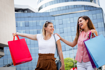 Wall Mural - Two smiling young woman walking down the street after sopping, carrying bags in their hands.