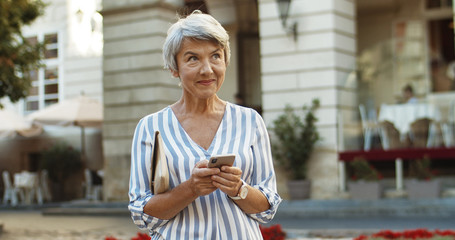 Caucasian beautiful old woman standing in center city and tapping message on smartphone. Grandmother using cellphone while typing sms. Senior nice female texting and scrolling on phone outdoors.