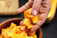 Pumpkin Pieces In Female Hand. Chopped Butternut Squash In A Wooden Bowl On Black Background, Closeup. Woman Throws Cutted Pumpkin Cubes Into A Salad Dish. Cooking Winter Squash, Food Ingredient