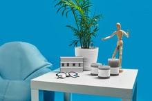 Turquoise Bean Bag, White Coffee Table With Green Flower In Pot, Three Candles, Wooden Figurine Of Human, Glasses. Blue Background. Close Up