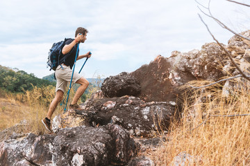 Youg traveler man climbing on stone with trekking poles and backpack walking on rock to the peak. Landscape trail travel concept.