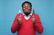 © Viktor Koldunov - Young hungry crazy african american man in red sweater holding a fork and a knife. Isolated on blue background