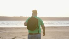A Plump Guy In A Green T-shirt Goes To The Water, Throwing A Backpack Over His Shoulder.