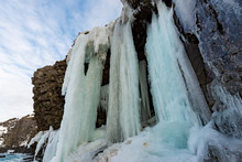 An Upward View Of A Wall Of Ice As The Water Falls Down Over The Rock Cliff. The Background Is Blue Sky With Thin White Clouds. The Icicles Spikes Are Both Yellow And Blue Hanging From A Rock Cliff.