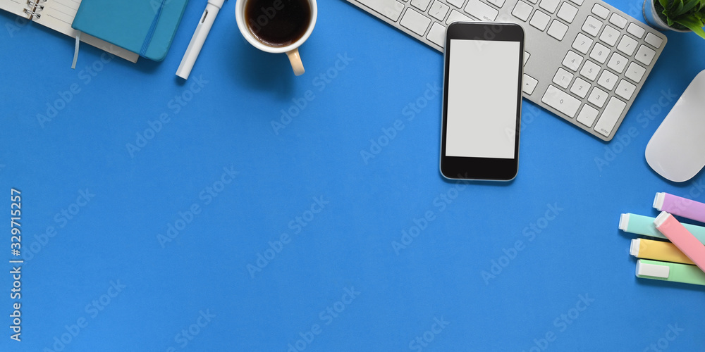 Top view image of blue working table with office equipment putting on it. Flat lay white blank screen mobile, wireless mouse and keyboard, marker pens, coffee cup, notebook, potted plant. - obrazy, fototapety, plakaty 