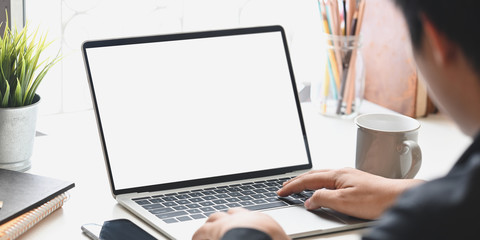 Cropped image of businessman typing on white blank screen computer laptop that putting on working desk with coffee cup, potted plant, pencil holder, books. Concentrated of work concept.