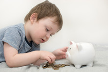 A Small, Cute Boy Lies And Puts Coins In A Piggy Bank. Piggy Bank In The Form Of A Pig. A Child Learns To Save Money. Close-up Photo, Cropped, On A Light Background.