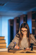 © bedya - Young student in glasses preparing for the exam. Girl in the evening sits at a table in the library with a pile of books