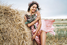 Young Brunette In A Pink Satin Dress, Without Linen Standing Near Hay Bales In Windy Weather. A Woman Holds A Bouquet Of Wildflowers
