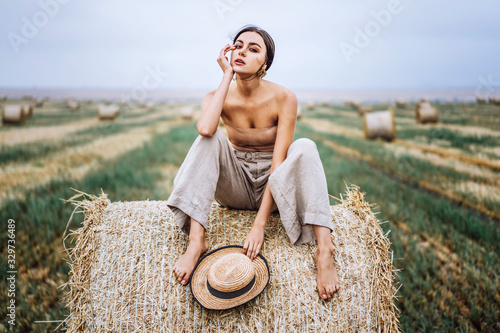 Fotomural Barefoot brunette in linen pants and bare shoulders sitting on a hay bales in warm autumn day