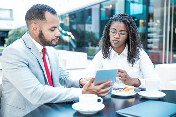 Wall Mural - Diverse business colleagues watching project presentation on tablet. Business man and woman sitting in cafe, using tablet together and talking. Media content concept