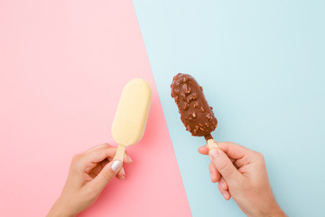 Young couple hands holding together ice creams with dark and white chocolate glaze. Closeup. Point of view shot. Light pink and blue table background. Pastel colors. Top down view.