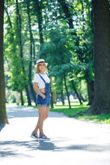 Wall Mural - young beautiful girl in denim overalls and a light hat walking in the park