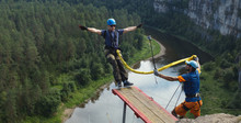 Young Man Jumping With A Rope From A Great Height Into The Deep Canyon On The Background Of The Forest And River Below, Panorama. Ropejumping.