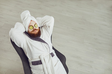 Funny fat bearded man with a cosmetic mask on his face in bathrobe towel on his head on his face resting against a gray background