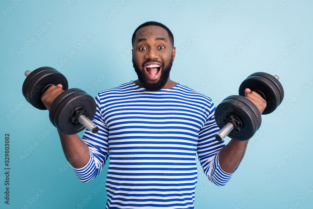 Photo of cool funny handsome dark skin guy open mouth lifting two heavy dumbbells weight practicing hard gym strong man wear striped sailor shirt isolated blue color background - obrazy, fototapety, plakaty 