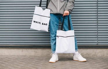 Closeup photo of bottom of stylish man in casual clothes holding 2 white eco bags in his hand. Advertising stylish reusable eco bags. Eco friendly guy wears stylish shopping bags