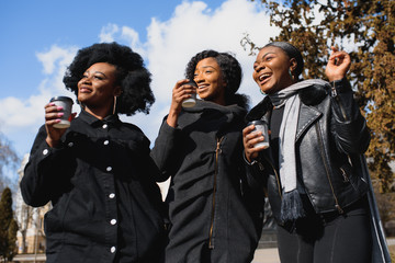 three beautiful and stylish dark-skinned girls with long hair standing in a city and drinking a coffee and use the phones