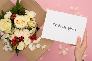 cropped view of woman holding thank you card near bouquet of flowers in cardboard box on pink background