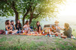 Picnic in the countryside - Group of young friends at sunset on spring day are sitting on the ground in a park near trees - They drinking red wine and eating grilled meat with barbecue and play guitar