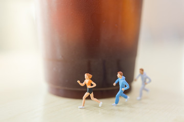 Wall Mural - Diet and food and Sport concept. Group of runner miniature figures people running on wooden table with takeaway plastic cup of iced black coffee (Americano).