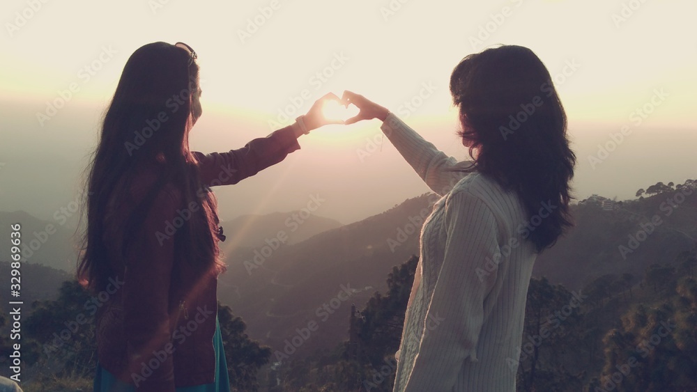 Female Friends Making Heart Shape From Hands In Front Of Sun Against Sky - obrazy, fototapety, plakaty 