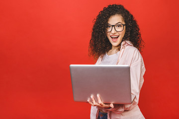 Wall Mural - Portrait of an excited curly young girl holding laptop computer isolated over red background.