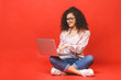 © denis_vermenko - Happy young curly beautiful woman sitting on the floor with crossed legs and using laptop on red background.