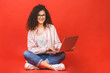 © denis_vermenko - Happy young curly beautiful woman sitting on the floor with crossed legs and using laptop on red background.