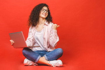 Happy young curly beautiful woman sitting on the floor with crossed legs and using laptop on red background.