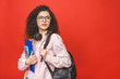 © denis_vermenko - Young curly student woman wearing backpack glasses holding books and tablet over isolated red background.